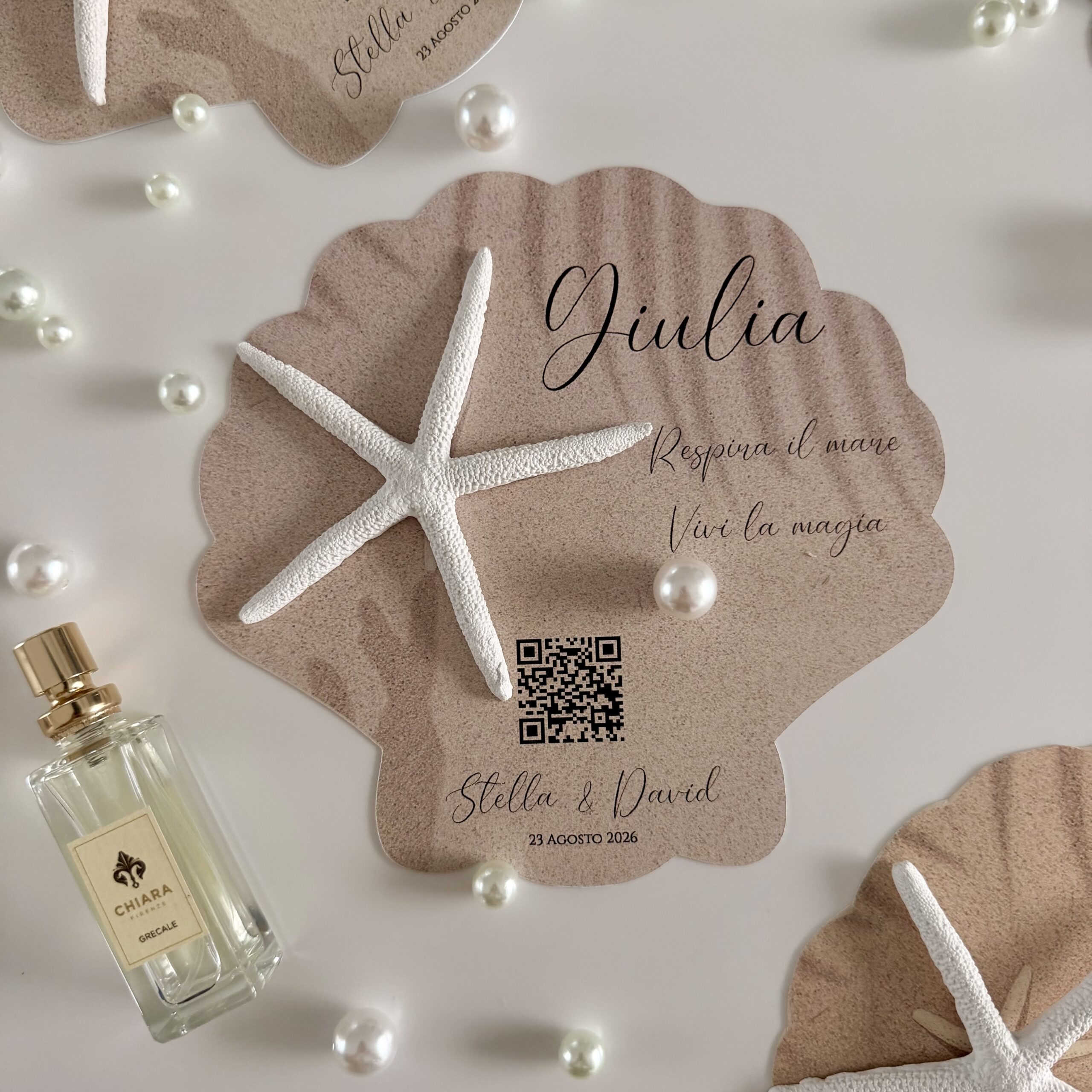 Segnaposto matrimonio a forma di conchiglia su sfondo bianco con perle e profumo, stella marina bianca applicata, personalizzazione completa con nome ospite in corsivo elegante, QR code album digitale e dettagli matrimonio